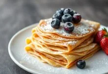 Pilha de panquecas douradas e finas, enroladas e empilhadas em um prato branco, com frutas ao lado.
