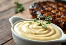 Molho Béarnaise cremoso e brilhante, com cor amarelo-ouro pálido, servido em uma tigela de cerâmica clara, acompanhado de folhas de estragão fresco e um bife grelhado ao fundo.