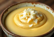 Tigela de sobremesa cremosa e amarelada, com textura lisa e brilhante, decorada com lascas de queijo.
