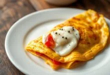 Omelete dourada e recheada, com muçarela derretida escapando pelo meio e fatias de tomate douradas, servida em prato branco sobre mesa de madeira.