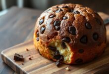 Um panettone de chocolate dourado e alto, fatiado sobre uma tábua de madeira, revelando o miolo macio e repleto de pedaços de chocolate derretido.