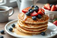 Pilha de panquecas douradas e fofas, cobertas com frutas vermelhas e um fio de mel.
