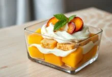 Uma travessa retangular de vidro com camadas visíveis de biscoito champagne amolecido, creme branco e aerado, e cubos de pêssego em calda dourados, finalizado com fatias de pêssego e folhas de hortelã.