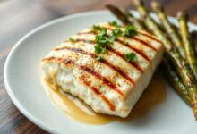 Filé de peixe branco grelhado com crosta dourada e levemente marcado pela grelha, coberto por um molho brilhante de manteiga derretida com limão e salsinha fresca, servido ao lado de legumes assados.