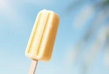 Um picolé de banana caseiro amarelo-claro, com textura cremosa e homogênea, perfeitamente desenformado em um palito branco, pronto para ser saboreado em um dia quente.