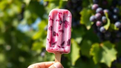 Um picolé de uva cremoso, de cor roxa intensa, com pedaços de uva visíveis, sendo segurado contra um fundo de verão.