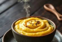Bowl de polenta cremosa amarela dourada, com textura aveludada e um fio de azeite brilhante, servida em prato fundo de cerâmica rústica.