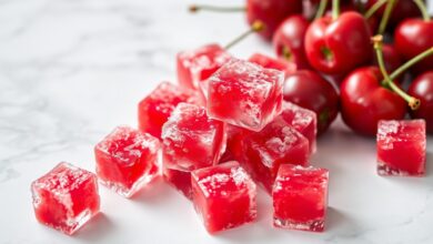 Cubos vibrantes de polpa de acerola caseira sobre uma superfície clara, prontos para o freezer, com acerolas frescas ao fundo.