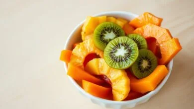 Tigela branca com fatias vibrantes de mamão papaia alaranjado, rodelas de abacaxi dourado e rodelas de kiwi verde com sementes pretas visíveis, dispostas de forma apetitosa sobre mesa de madeira clara.