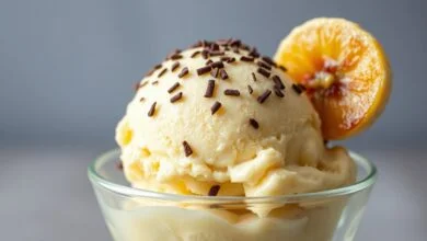 Taça de vidro transparente com uma bola generosa de sorvete de banana de cor amarela pálida, finalizado com gotas de chocolate e uma rodela de banana caramelizada na borda.