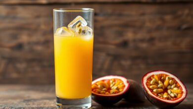 Um copo alto de suco de maracujá vibrante e amarelo-alaranjado, com alguns grãos flutuando e cubos de gelo, servido sobre uma superfície rústica.
