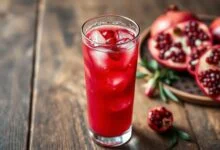 Um copo alto de suco de romã de cor vibrante e intensa, servido com gelo e alguns grãos de romã flutuando, pronto para ser saboreado.