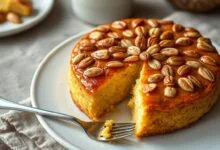Torta redonda com topo dourado e brilhante de amêndoas caramelizadas, base macia visível na fatia, apresentada em prato de porcelana branca