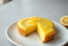 Uma torta de limão francesa inteira, com recheio de creme de limão liso e brilhante, cor amarelo-pálido, sobre uma base de massa dourada. Decorada discretamente com raspas de limão siciliano, fatiada para revelar a textura sedosa do interior.
