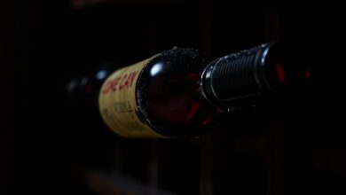 Garrafas de vinho tinto deitadas em uma adega de madeira rústica e escura, com uma poeira sutil indicando o tempo, iluminadas por uma luz suave que destaca a cor rubi profunda do vinho no interior.