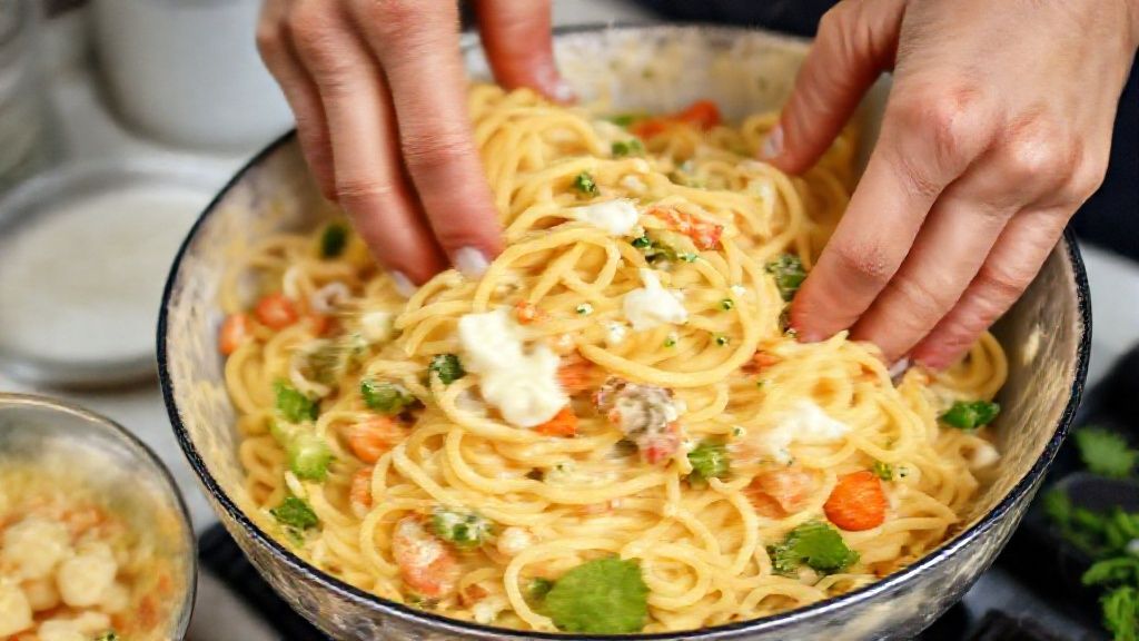 Mãos misturando o macarrão cozido com legumes coloridos e maionese em uma tigela