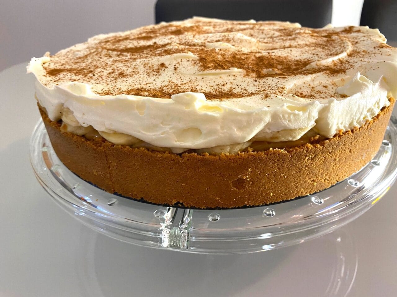 Fatia de banoffee em um prato, mostrando as camadas de biscoito, doce de leite, banana e chantilly, polvilhada com cacau em pó.